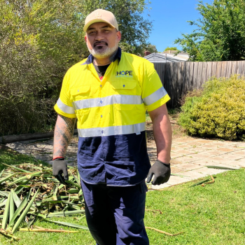 A smiling man in a Hope Industries yellow high-visibility shirt and cap standing in a garden beside a pile of cleared vegetation.